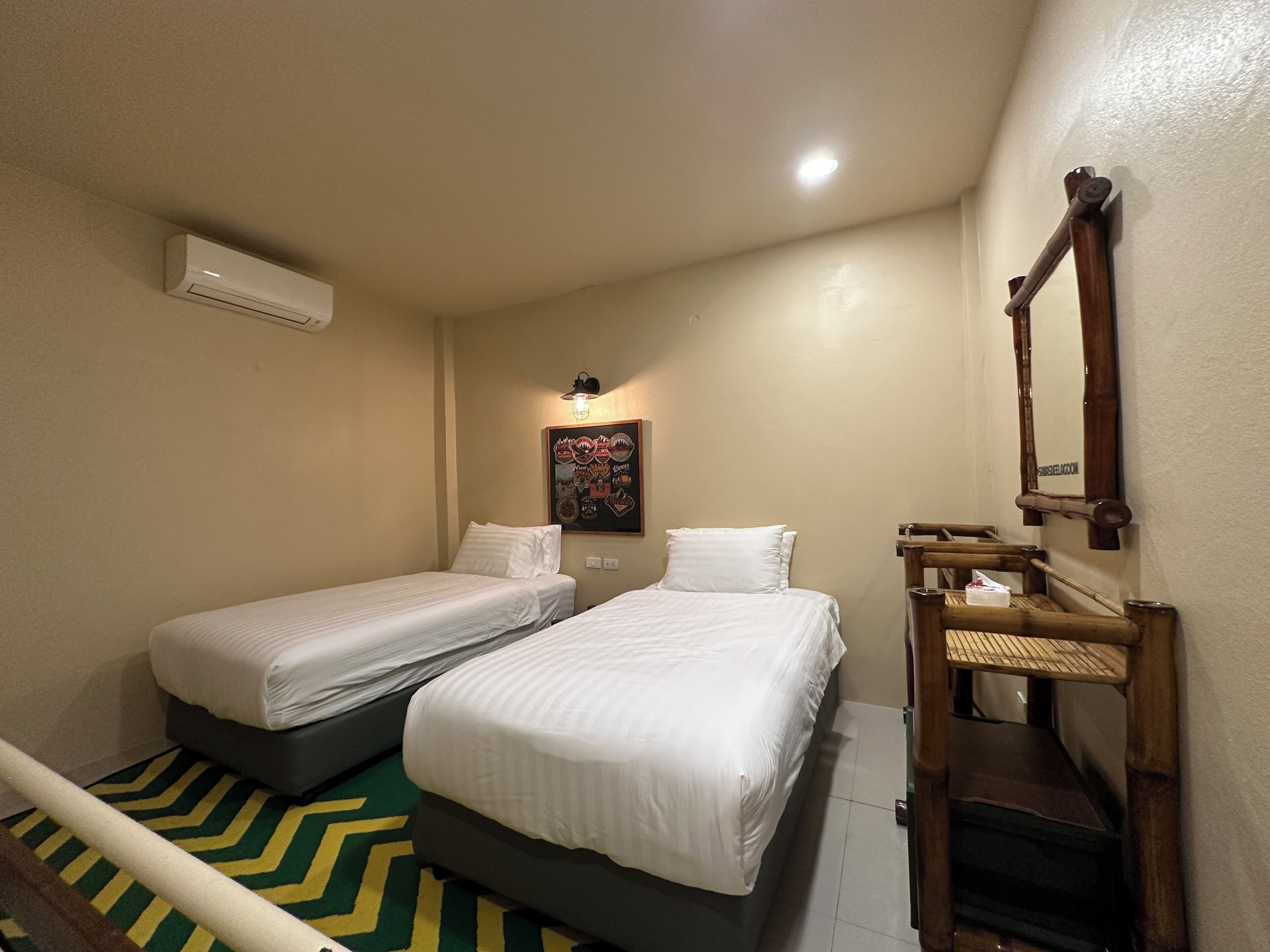 Une petite chambre d’hôtel avec deux lits simples, un banc en bois, un miroir fixé au mur et un climatiseur fixé au mur.