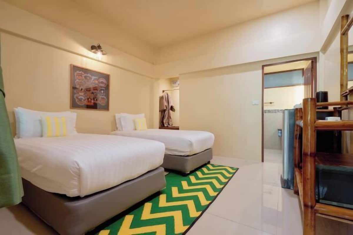 Een hotelkamer met twee bedden, een vloerkleed met een groen en geel patroon, een houten stoel en een schilderij aan de muur.