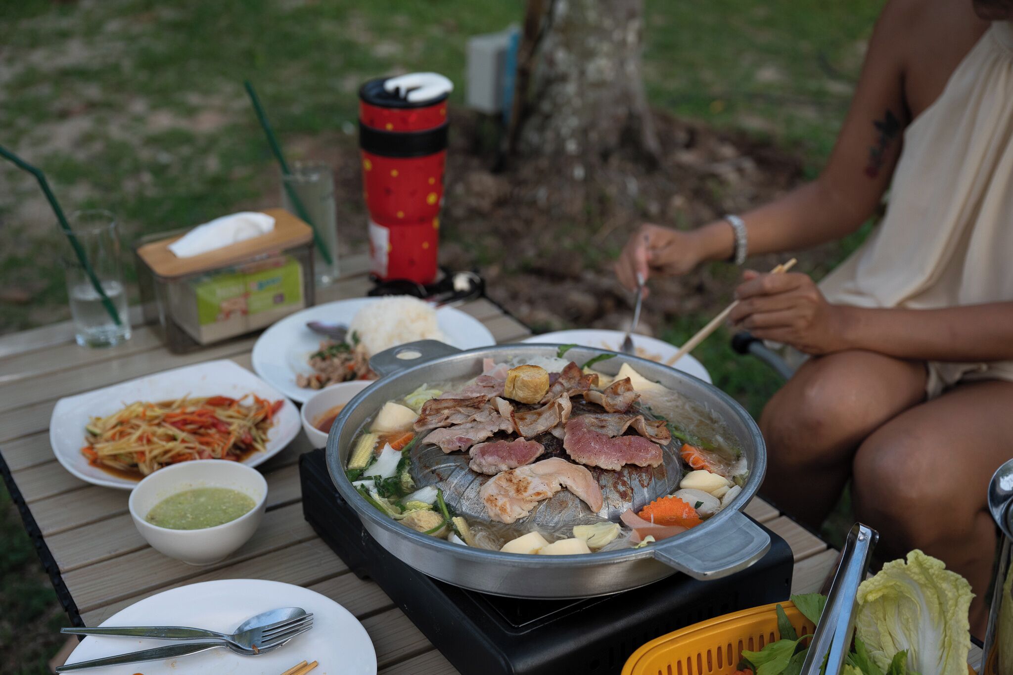 Una mesa de comedor con un recipiente de cocción caliente, platos de comida, un termo y una cesta.
