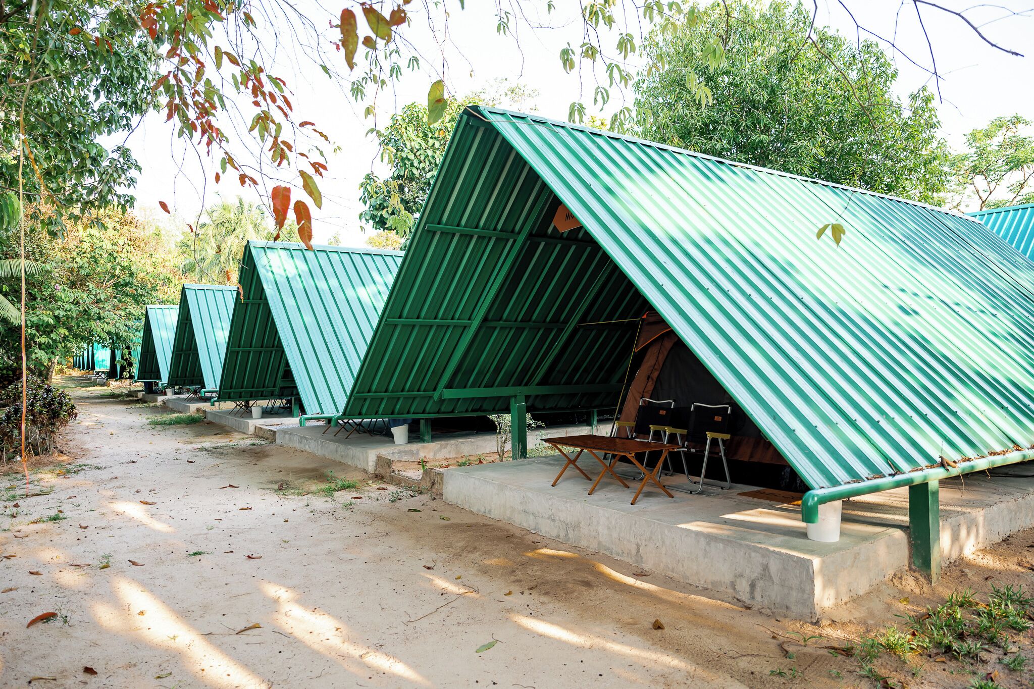 A row of green-roofed, triangular tents with concrete steps leading up to them, set in a natural environment with trees and a dirt path.