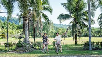 Two people on bicycles with baskets, surrounded by palm trees and greenery.