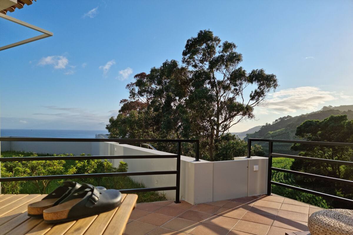 Ein Balkon mit Blick auf das Meer, einen Baum und ein Paar Sandalen auf einem Holztisch.