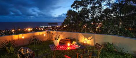 An outdoor dining area with a table and chairs, surrounded by greenery and a view of the city at dusk.