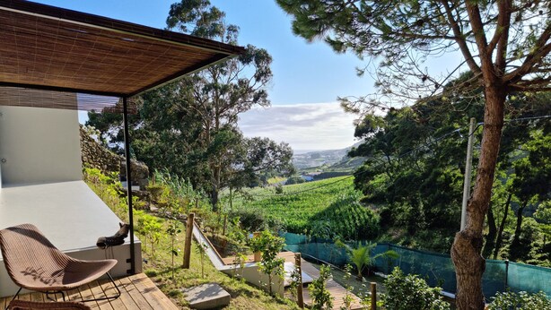 A terrace with wicker chairs, a wooden deck, and a view of a valley with a river. - Place Of Moments Natur (Vila Franca do Campo)