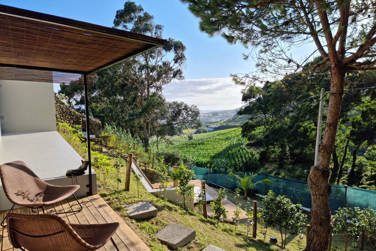 Eine Terrasse mit Korbstühlen, einer Holzdielenfläche und Blick auf ein Tal mit einem Fluss.