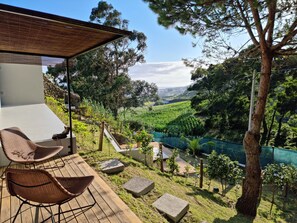 A terrace with wicker chairs, a wooden deck, and a view of a valley with a river. - Place Of Moments Natur (Vila Franca do Campo)