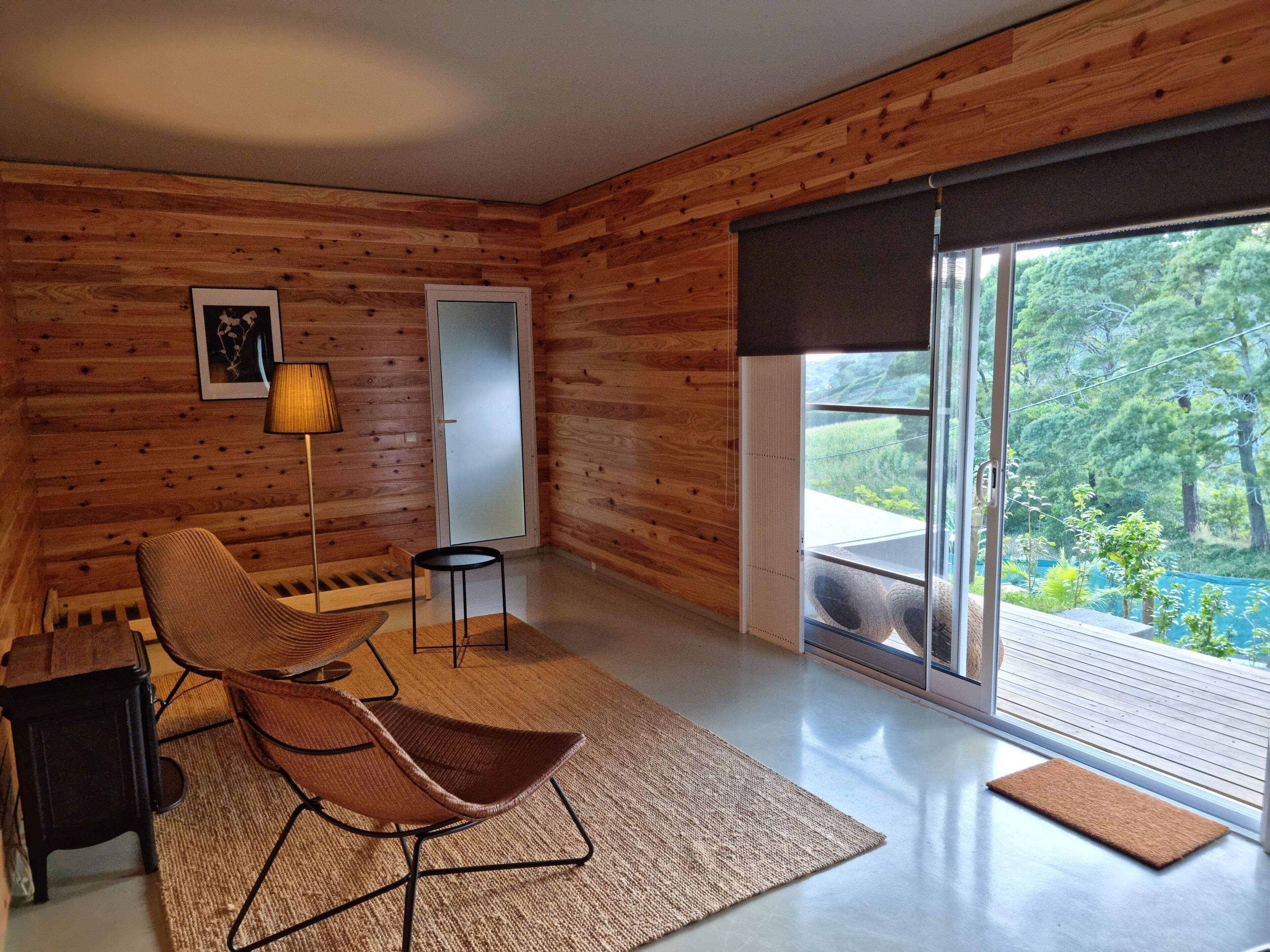 Een moderne kamer met houten wanden, een grote schuifglazen deur naar een balkon, een stoel, een tafeltje en een lamp.