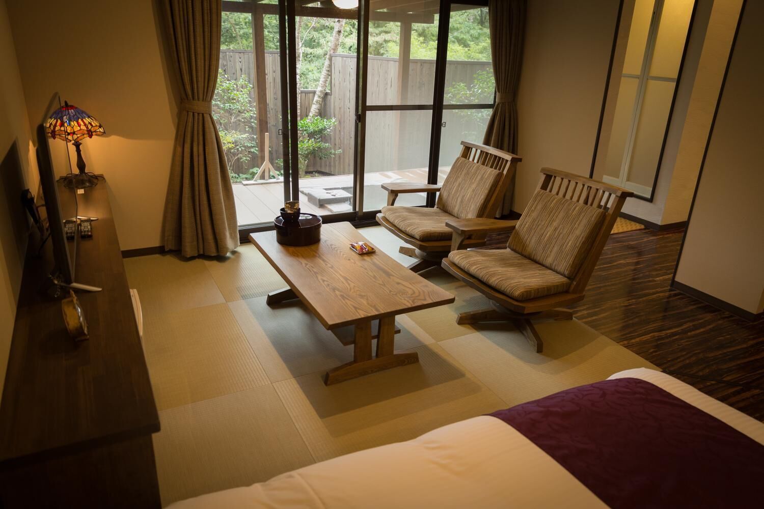 A hotel room with a bed, a wooden table, two chairs, a TV, and a window with a view of the outdoors.