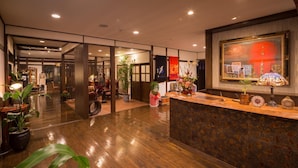 A hotel lobby with a wooden reception desk, potted plants, and a large painting on the wall.
