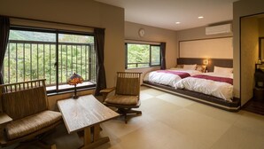 A hotel room with a bed, two chairs, a table, and large windows offering a view of greenery.