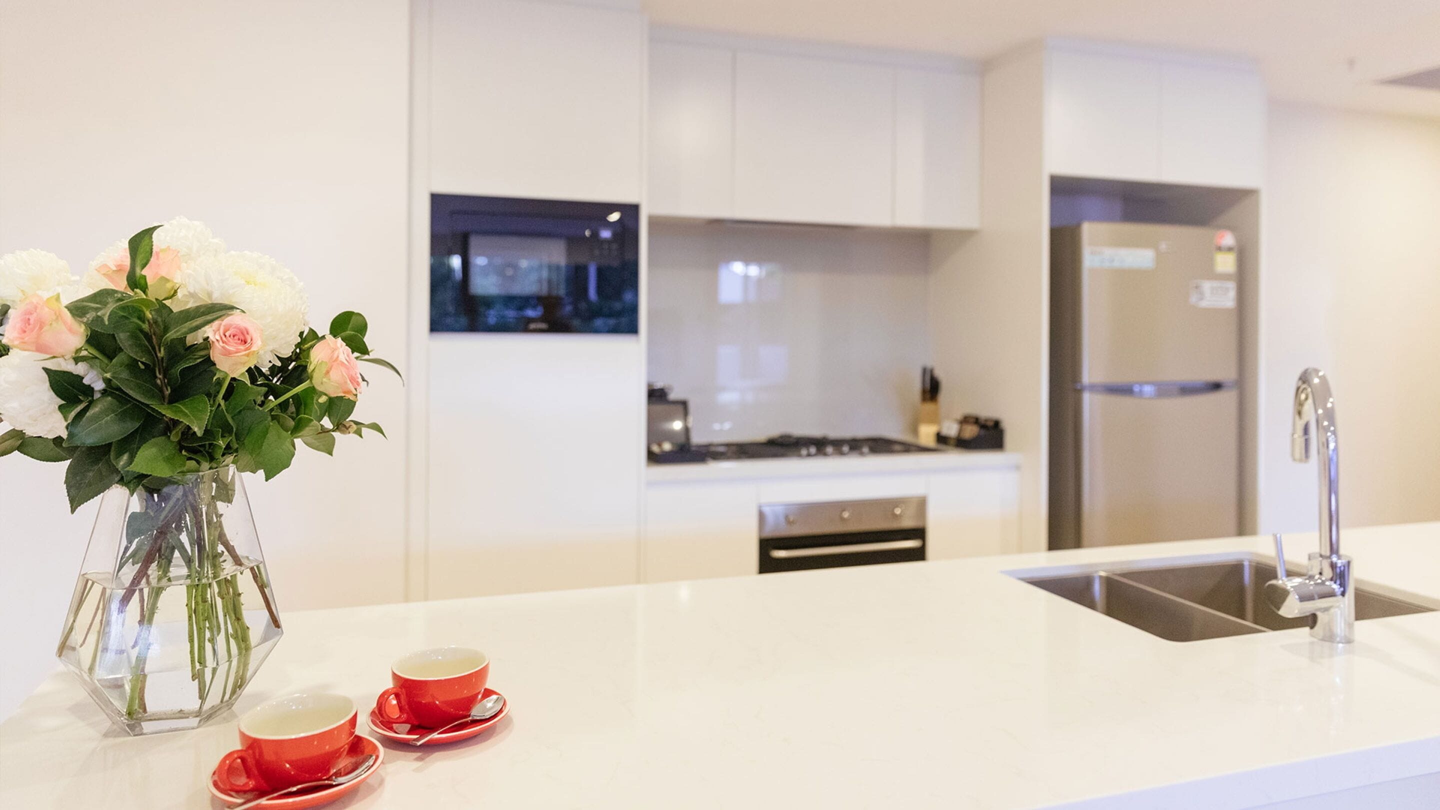Une cuisine moderne avec des meubles blancs, un évier en acier inoxydable et un vase rempli de fleurs posé sur le plan de travail.