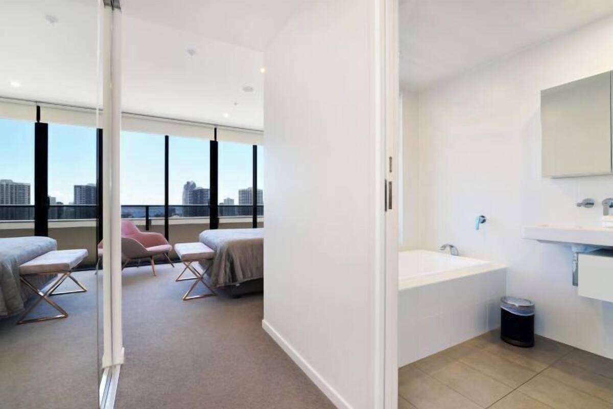 A modern hotel room with a large window, a bathtub, a sink, and a chair.