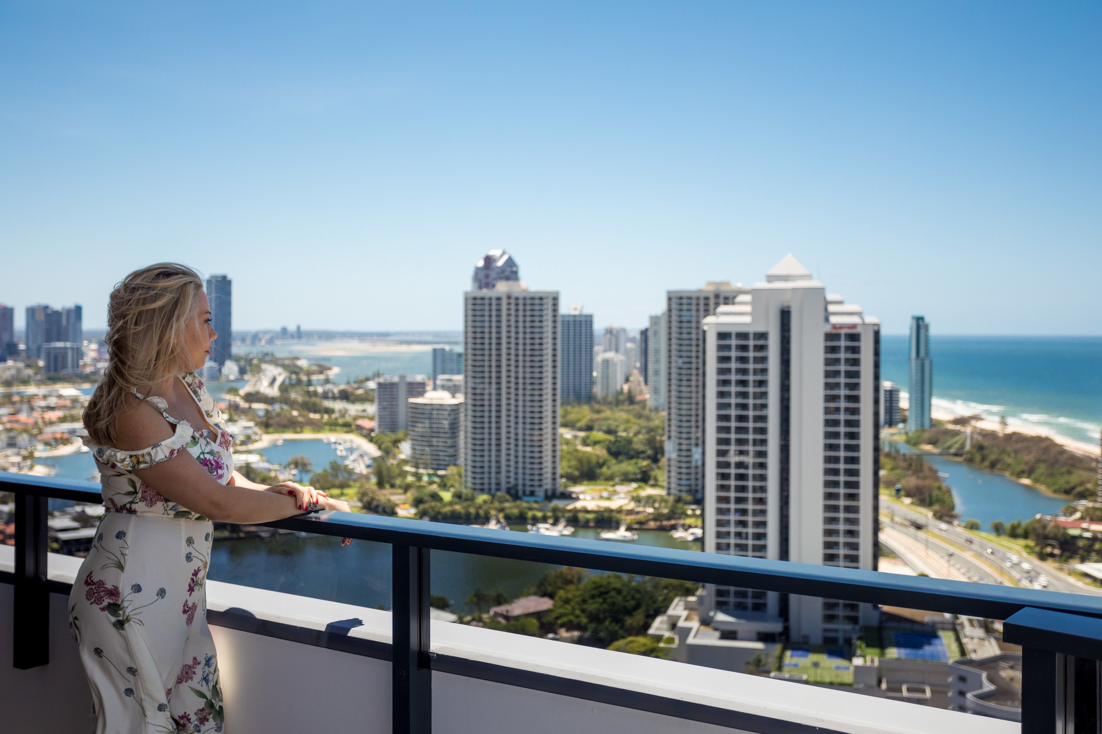 Una mujer de pie junto a un balcón con vista a un paisaje urbano con edificios altos y una playa.