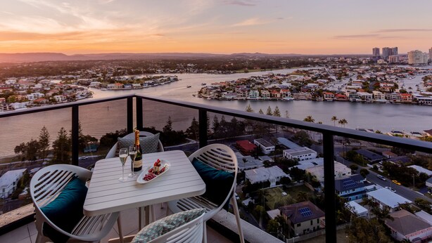 A balcony with a table set for two, overlooking a river and cityscape at sunset. - Ruby Gold Coast by CLLIX (Surfers Paradise)