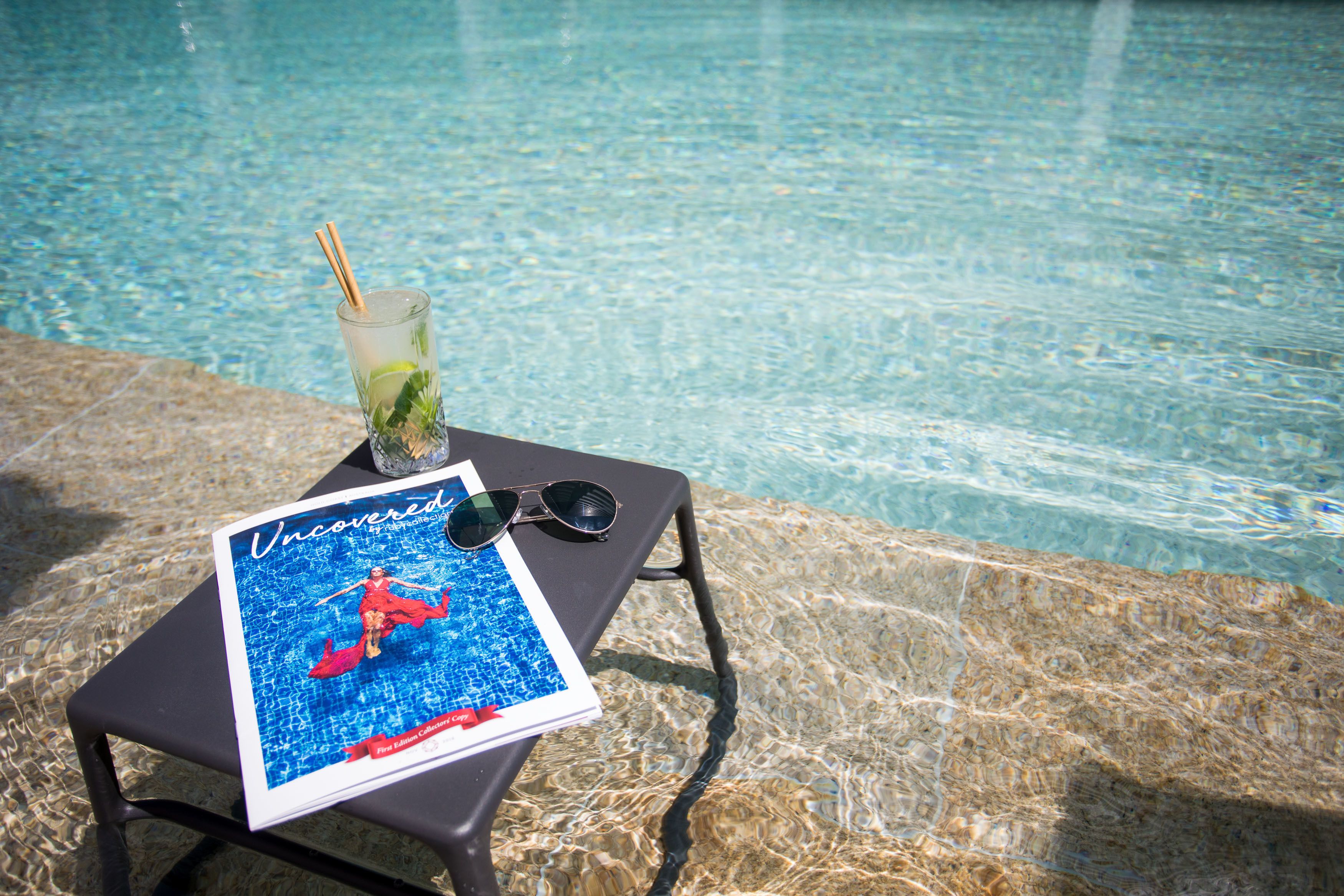 A glass of drink with a straw and mint leaves on a table by a pool.