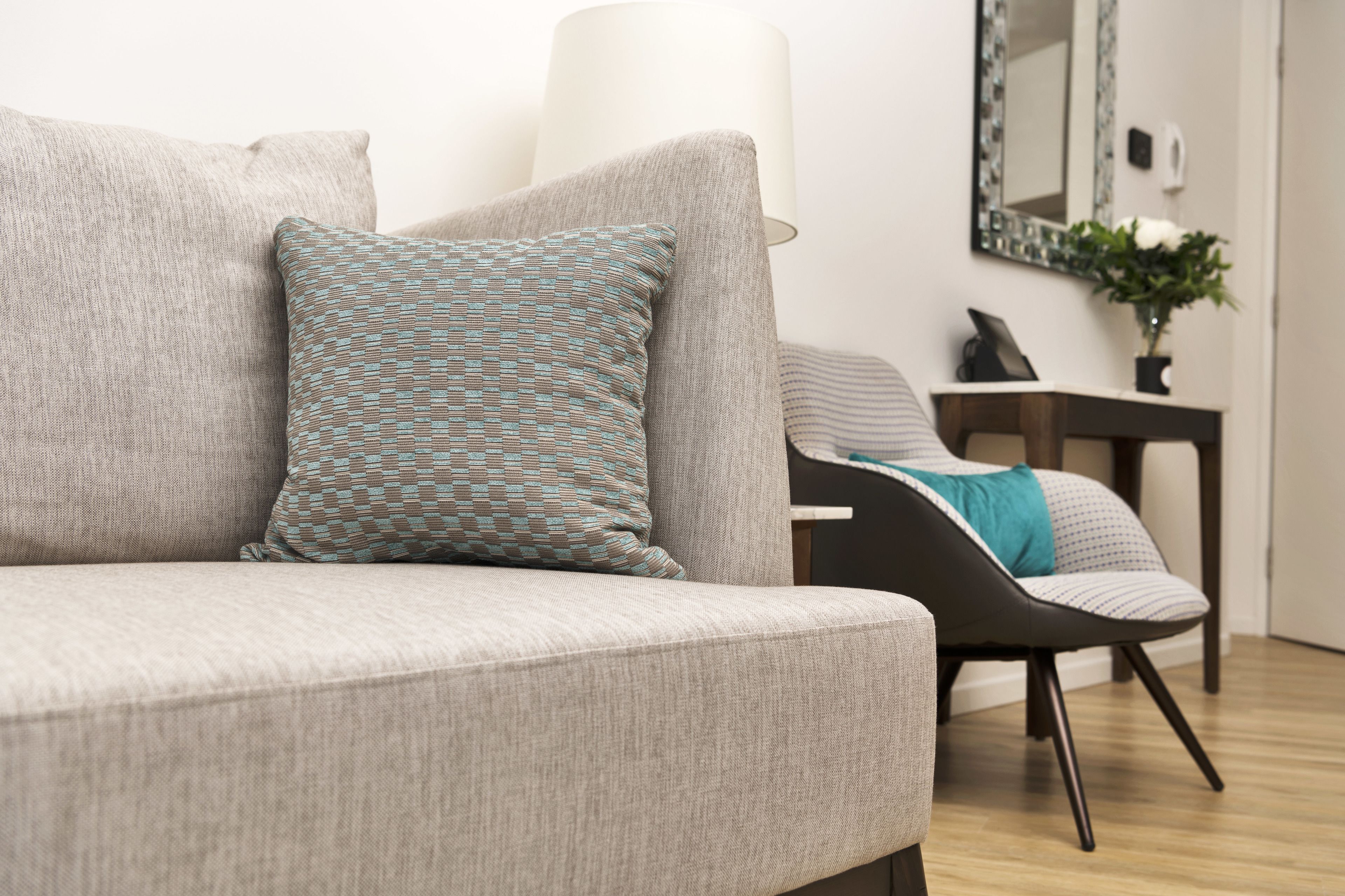 A modern living room with a grey sofa, patterned pillows, a black chair, and a small table with a lamp and a vase of flowers.