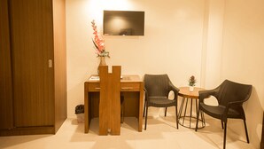 A small, well-lit room with a wooden desk, two chairs, a TV mounted on the wall, and a vase with flowers on the desk. - Rizmy Hotel (Cabuyao)