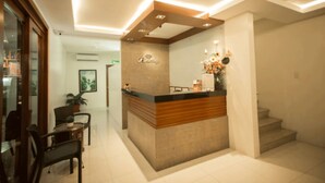A reception desk with a wooden counter and a sign above it. There is a staircase to the right and a chair and table in the foreground. - Rizmy Hotel (Cabuyao)