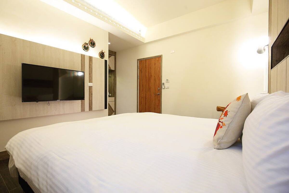 A hotel room with a large bed, a flat-screen TV mounted on the wall, a wooden door, and a small table with a lamp.