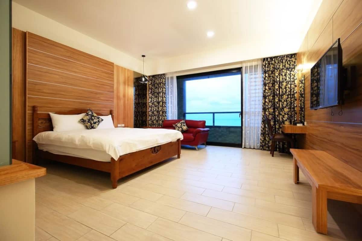 A modern bedroom with a wooden bed, a red armchair, a wooden bench, and a large window with curtains.