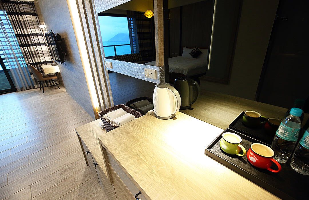 Quarto de hotel moderno com piso de madeira, área de jantar com mesa e cadeiras, cozinha com chaleira e xícaras, e uma cama com vista para o mar.