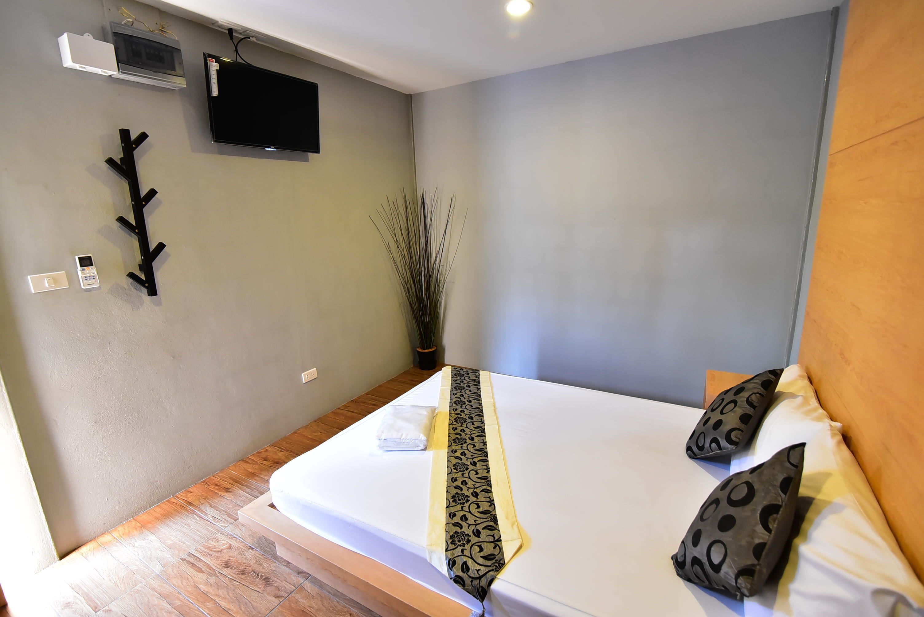 A neatly made bed with a patterned runner, two pillows with a circular design, a wall-mounted TV, a small plant, and a wall-mounted air conditioning unit.