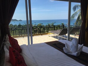 Habitació d'hotel amb llit, finestra amb vista a l'oceà i embarcacions, balcó amb cadira i un ambient tropical.