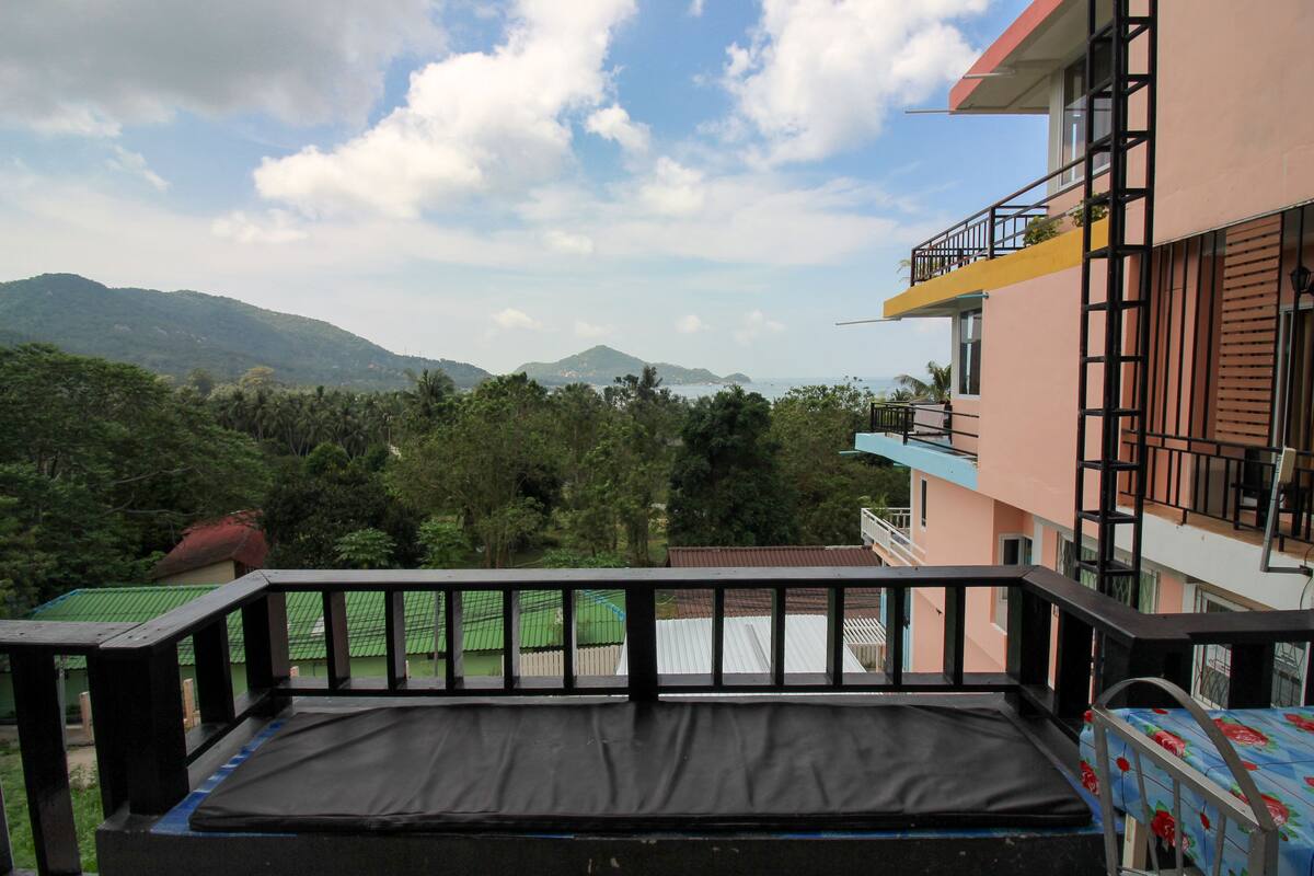 Ein Balkon mit schwarzer Matratze, mit Blick auf eine gebirgige Landschaft und ein Gebäude mit Balkonen.