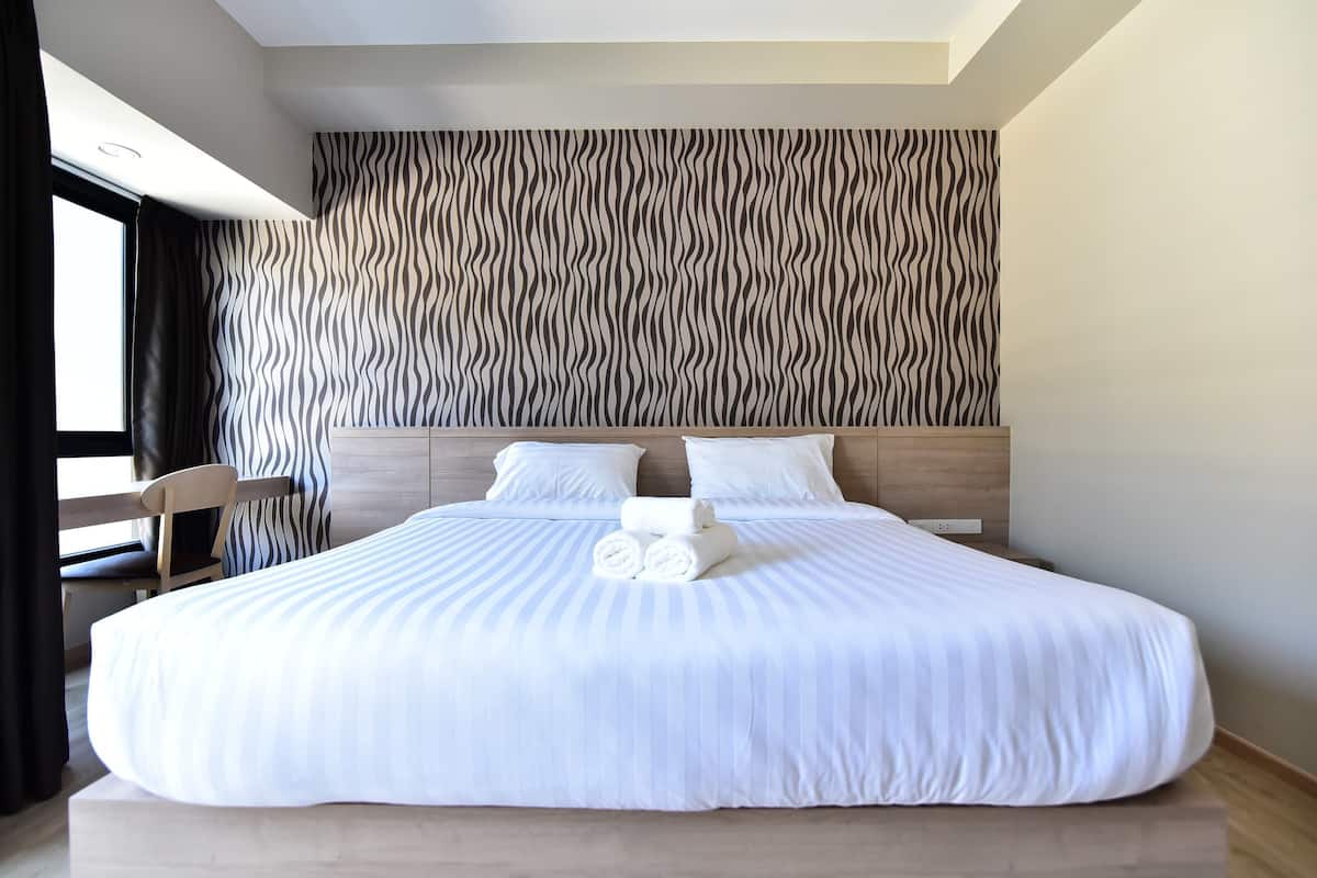 Un grand lit recouvert de draps blancs, deux oreillers et des serviettes enroulées dans une chambre d’hôtel moderne, avec un mur à motifs ondulés et une tête de lit en bois.