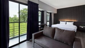A modern hotel room with a large window, a sofa, a bed with white linens, and a dark headboard. - At S115 Residence (Samut Prakan)