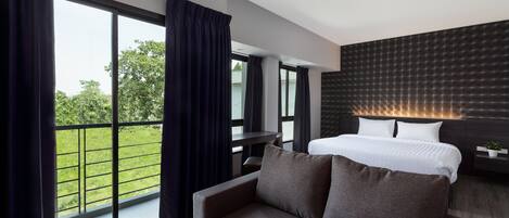 Une chambre d’hôtel moderne dotée d’une grande fenêtre, d’un canapé, d’un lit avec du linge de lit blanc et d’une tête de lit sombre.