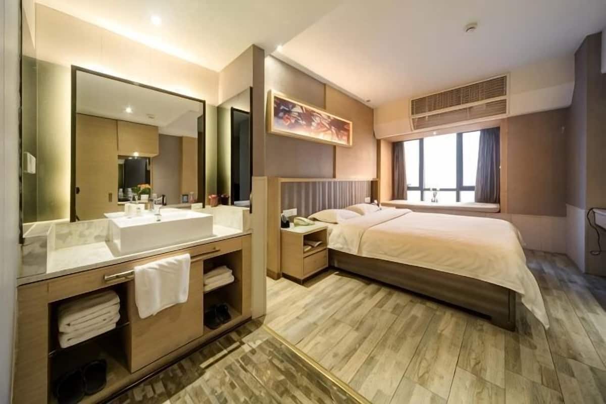 A hotel room with a large bed, a bedside table, a nightstand, a bathroom with a sink and mirror, and a window providing natural light.