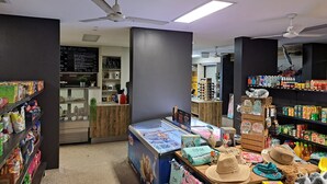 Interior de una tienda de comestibles con estantes llenos de refrigerios, una vitrina refrigerada y una caja registradora.