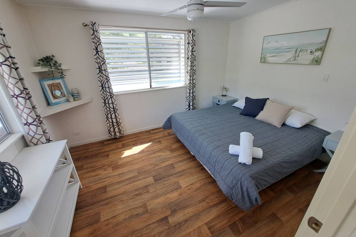 Une chambre à coucher avec un lit, une table de chevet, une fenêtre avec des stores, un ventilateur de plafond et un tableau accroché au mur.