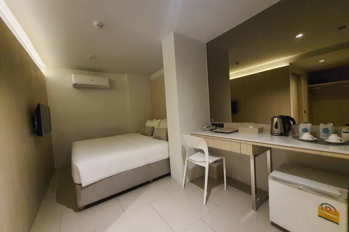 Habitación de hotel compacta con una cama individual, un escritorio con silla, un refrigerador pequeño y un espejo.