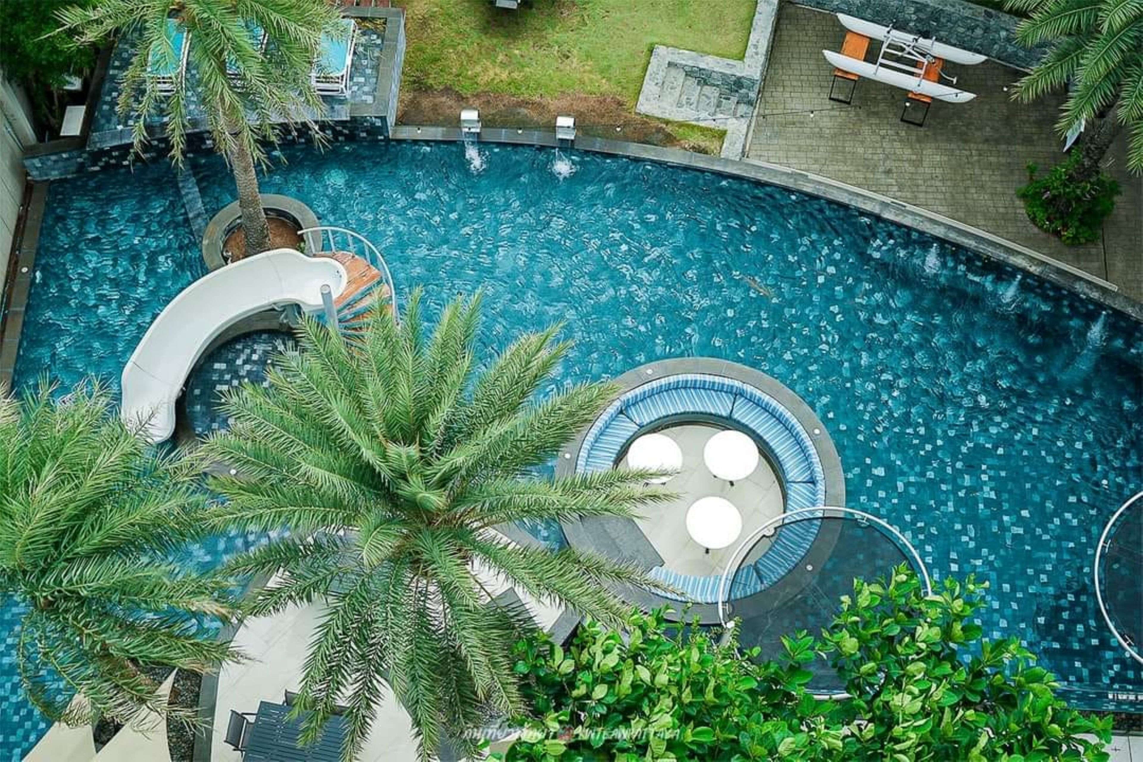 A large, curved swimming pool with a slide, surrounded by palm trees and a small circular seating area.