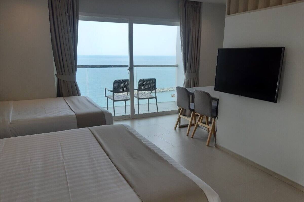 Habitación de hotel con dos camas, televisor, escritorio con silla y balcón con vista al mar.