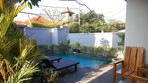 A swimming pool with lounge chairs, surrounded by a low wall and greenery. - Sweet Corner Guest House (Denpasar)