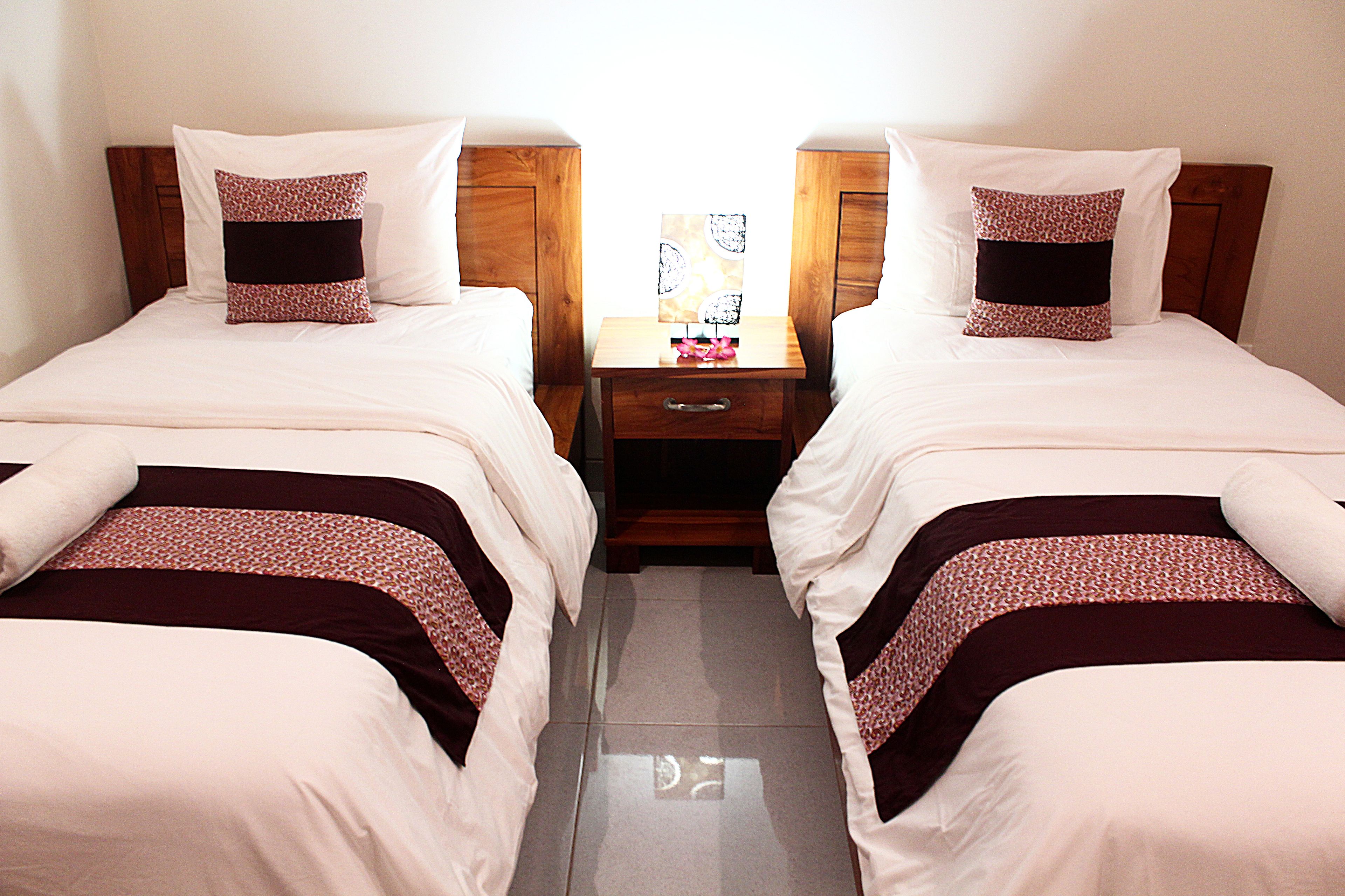 A hotel room with two single beds, each with a wooden headboard, white bedding, and a patterned pillow. There is a wooden nightstand between the beds with a small table and a lamp.