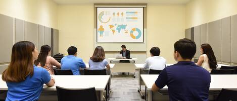 A conference room with a presentation screen, white tables, and black chairs.