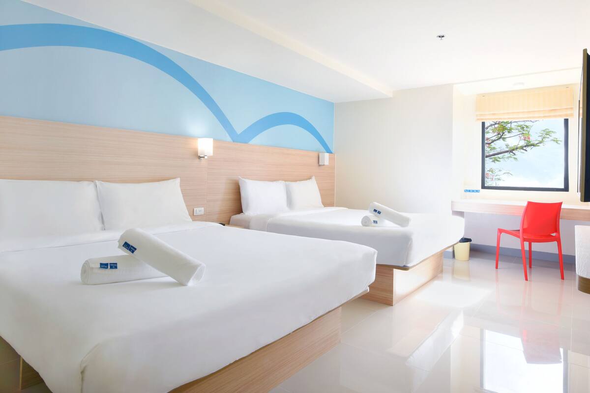 A hotel room with two beds, a wooden headboard, a red chair, and a window with a view of trees.