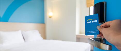 Una mano insertando una tarjeta en una cerradura digital en una puerta azul, con una cama y una lámpara de pared visibles al fondo.