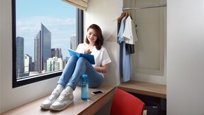 A person sitting by a window with a city view, a desk, a red chair, and a clothing rack with hangers. - Hop Inn Hotel Alabang Manila (Muntinlupa)