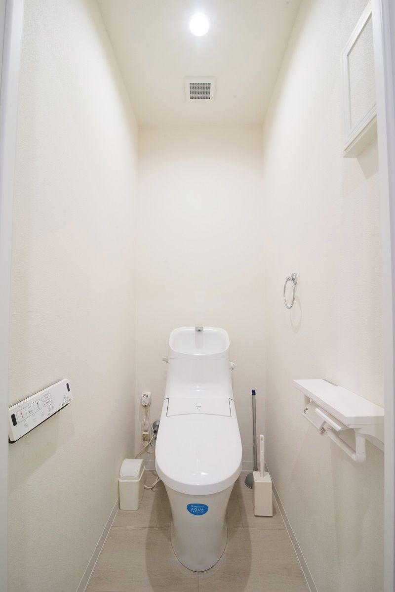 A small, white, single-occupancy bathroom with a toilet, a small trash bin, a wall-mounted shelf, and a power outlet.