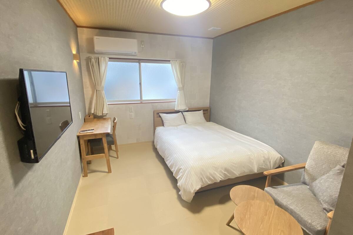 A hotel room with a bed, a desk with a chair, a TV, and a window with curtains.