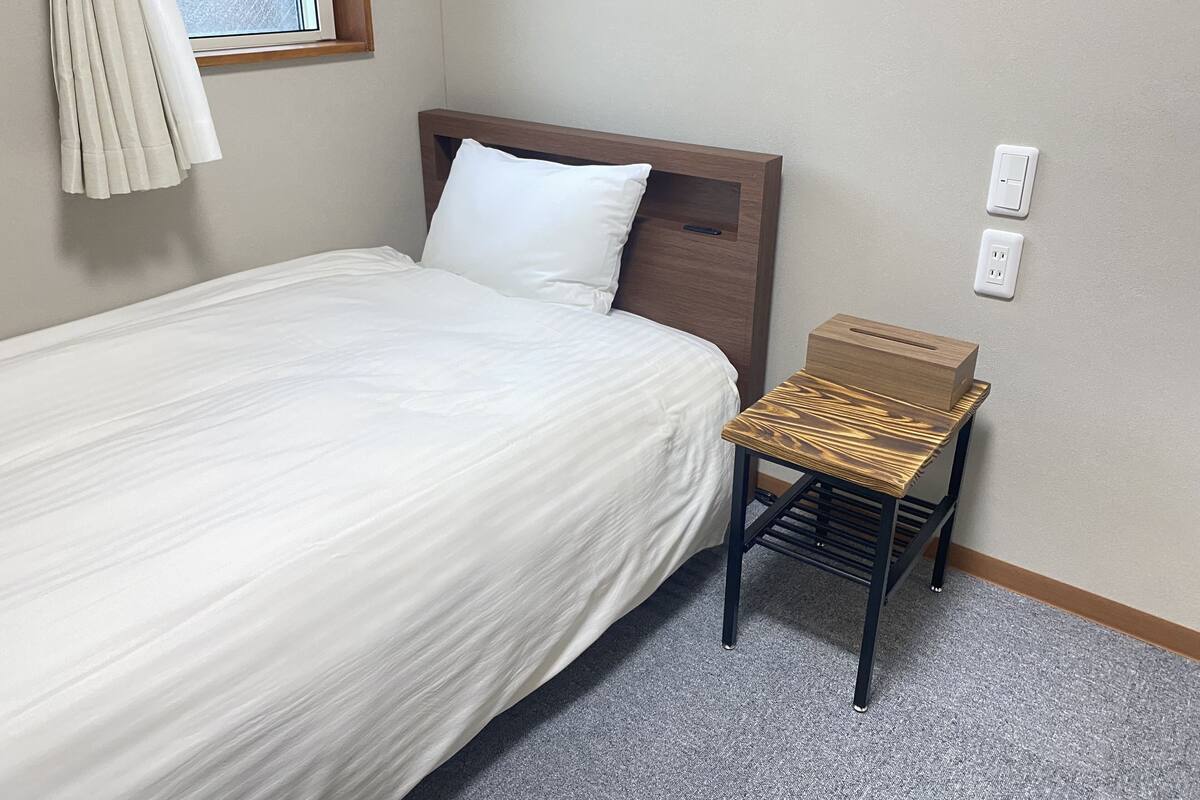 A neatly made bed with a wooden headboard, a small wooden side table, and a window with curtains.