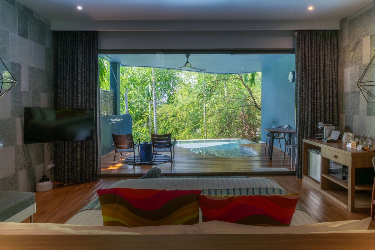 A modern bedroom with a large bed, a TV, a desk, and a view of a lush garden.