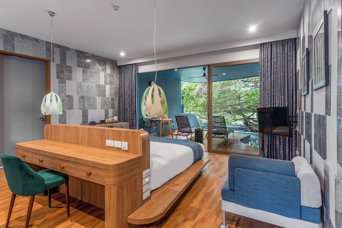 A modern hotel room with a wooden desk, a bed, a blue sofa, and a balcony with a pool.