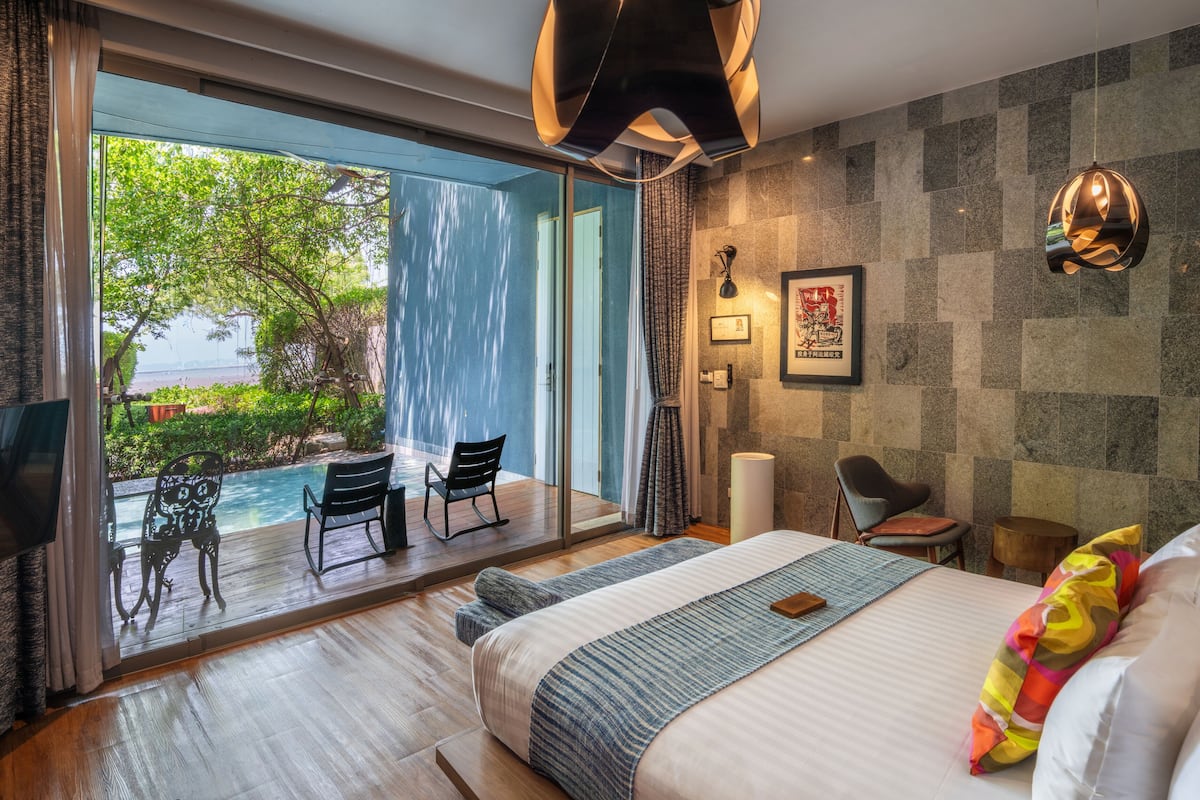 A modern hotel room with a large bed, a desk, a chair, a TV, and a view of a pool and greenery.