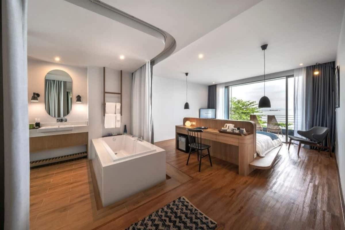 A modern hotel room with a large bathtub, a desk with a chair, a bed, and a large window with a view.